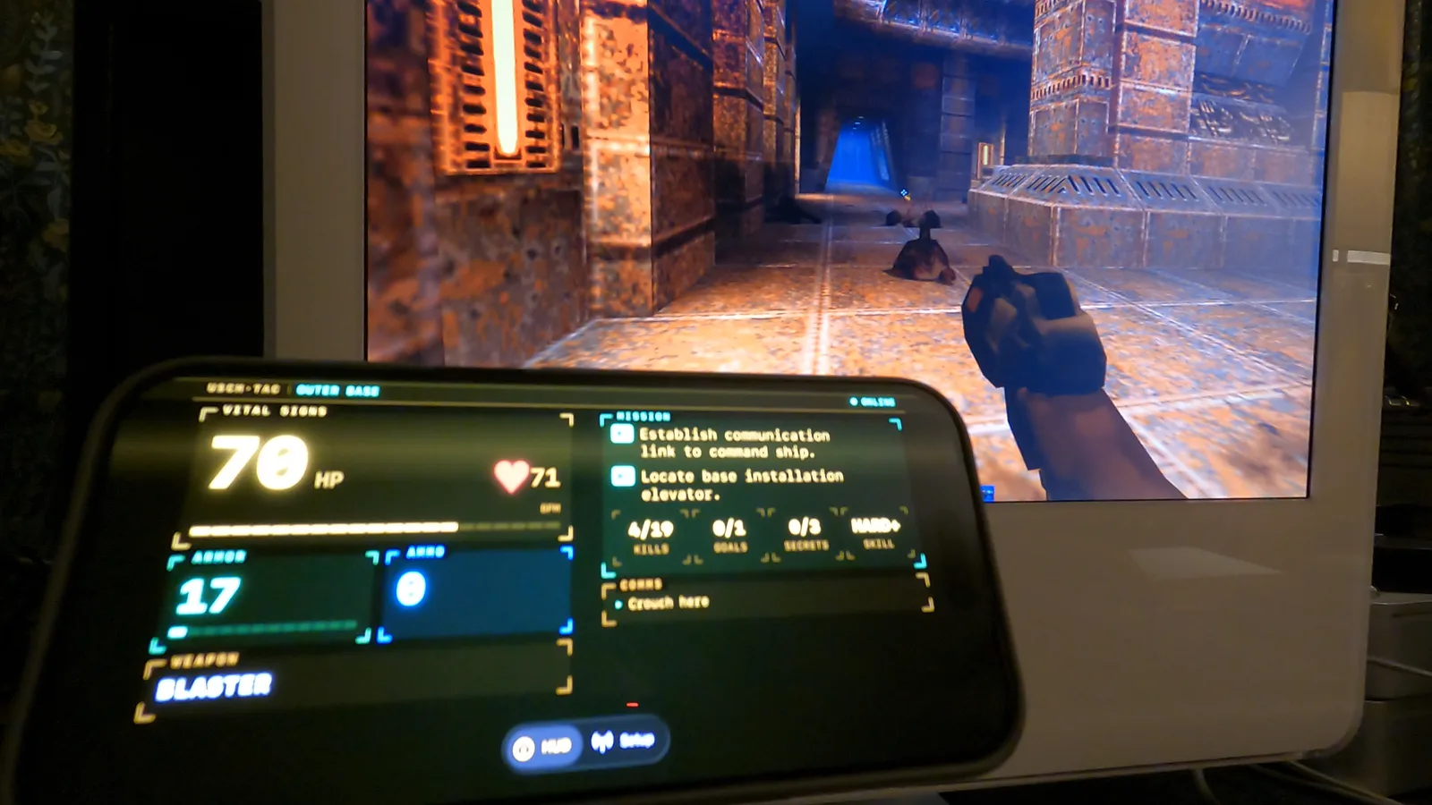 A 2005 iMac G5 running Quake II in a corridor, with an iPhone on a stand beside it showing the tactical computer HUD at 70 HP